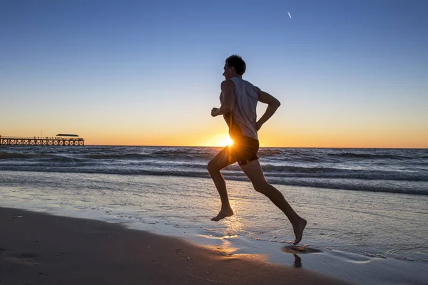 Siluet atlet muda yang dinamis pelari manusia dengan pelatihan tubuh yang cocok pada musim panas matahari terbenam pantai berjalan bertelanjang kaki dalam olahraga yang sehat dan kebugaran konsep — Stok Foto