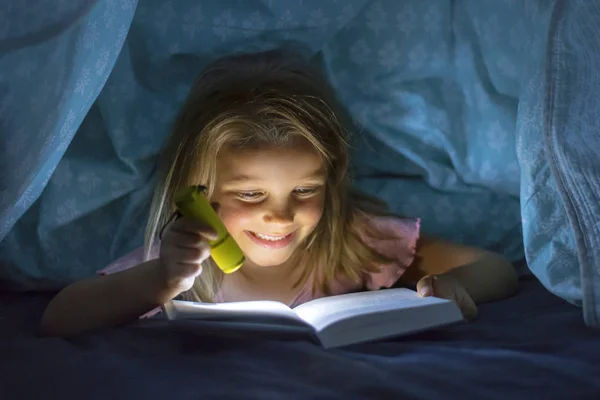 Сладкий красивый и красивый маленький блондинка девочка 6 до 8 лет под одеялом чтение книги в темноте ночью с факелом свет улыбаясь счастливым — стоковое фото