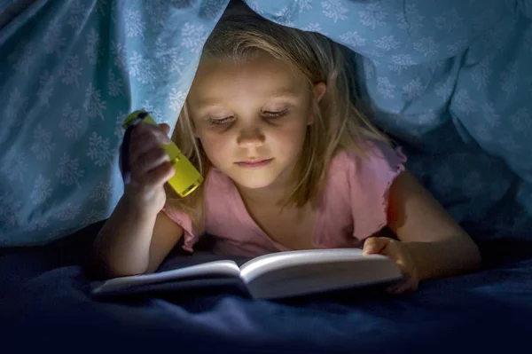 Солодка красива і досить маленька блондинка 6-8 років під ліжком обкладинки читання книги в темряві вночі з факелом світла посміхається щасливий — стокове фото