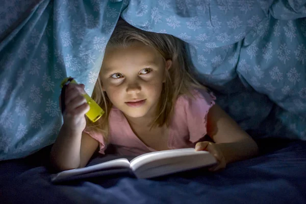 Doce linda e bonita menina loira 6 a 8 anos de idade sob a cama cobre livro de leitura no escuro à noite com luz da tocha sorrindo feliz — Fotografia de Stock