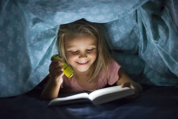 Солодка красива і досить маленька блондинка 6-8 років під ліжком обкладинки читання книги в темряві вночі з факелом світла посміхається щасливий — стокове фото