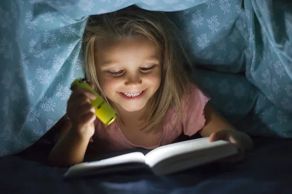 Сладкий красивый и красивый маленький блондинка девочка 6 до 8 лет под одеялом чтение книги в темноте ночью с факелом свет улыбаясь счастливым — стоковое фото