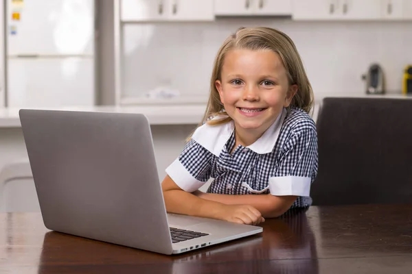तरुण सुंदर आणि गोड लहान मुलगी 6 ते 8 वर्षांची गोरी केस आणि निळे डोळे घरी स्वयंपाकघरात बसून लॅपटॉप संगणकासह आनंदी हसत — स्टॉक फोटो, इमेज