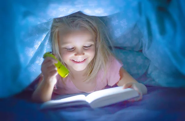 Sweet beautiful and pretty little blond child girl 6 to 8 years old lying under bed covers reading book in the dark with torch flashlight smiling happy — Stock Photo, Image