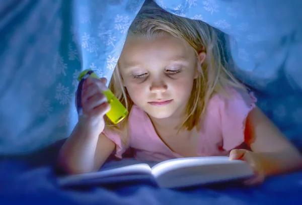 Мила красива і досить маленька блондинка дівчинка 6-8 років лежить під ліжком обкладинки читання книги в темряві з факелом ліхтарик посміхається щасливий — стокове фото