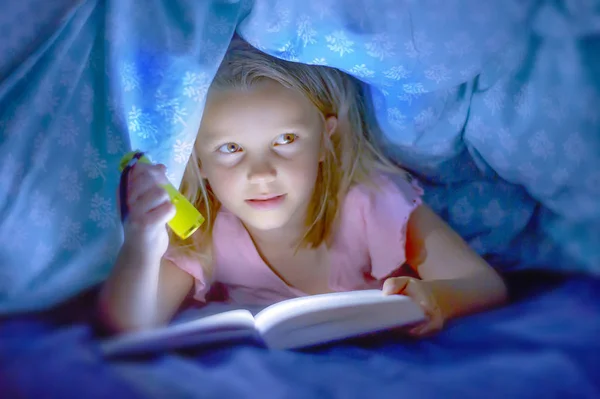 Sweet beautiful and pretty little blond child girl 6 to 8 years old lying under bed covers reading book in the dark with torch flashlight smiling happy — Stock Photo, Image