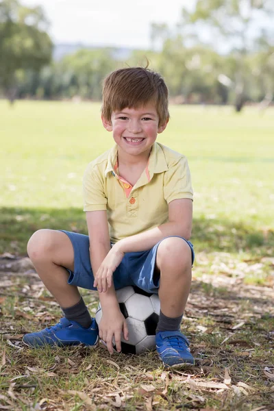 Kid 7 or 8 years old enjoying happy playing football soccer at grass city park field posing smiling proud sitting on the ball in childhood sport passion — Stock Photo, Image