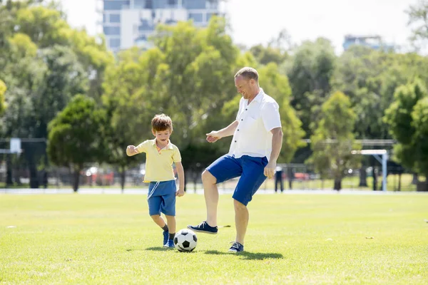 Молодой счастливый отец и взволнованный маленький 7 или 8-летний сын играют вместе футбол в саду городского парка работает на траве ногами мяч — стоковое фото