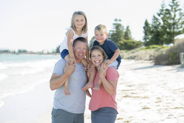 Jovem feliz bela família ter férias na praia sorrindo mãe e pai carregando pequeno filho e filha jovem nas costas — Fotografia de Stock