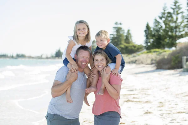Joven feliz hermosa familia tener vacaciones en la playa sonriente mamá y papá llevando pequeño hijo y joven hija en la espalda — Foto de Stock