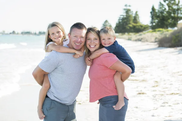 ママとパパの幼い息子と娘を背負って笑顔のビーチでの休日を持つ若い幸せな美しい家族 — ストック写真