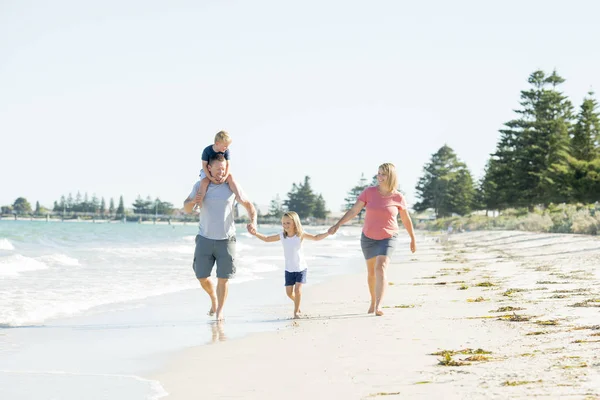 युवा खुश और सुंदर परिवार माँ पिता बेटे और बेटी का हाथ पकड़ते हुए समुद्र तट पर खुश चलते हुए गर्मियों की छुट्टियों का आनंद लेते हुए — स्टॉक फ़ोटो, इमेज