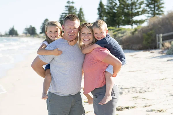 Young happy beautiful family having holidays on beach smiling mom and dad carrying little son and young daughter on the back — Stock Photo, Image