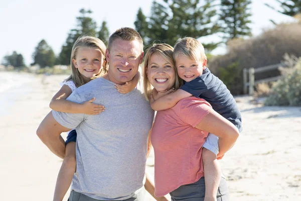 Junge glückliche schöne Familie macht Urlaub am Strand lächelnd Mama und Papa tragen kleinen Sohn und kleine Tochter auf dem Rücken — Stockfoto