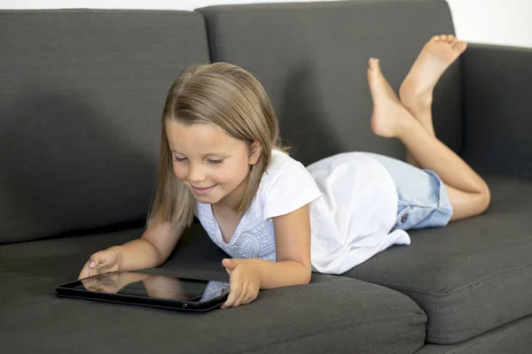 Giovane dolce e felice bambina 6 o 7 anni sdraiata sul divano del salotto di casa utilizzando internet tablet digitale touch pad — Foto Stock