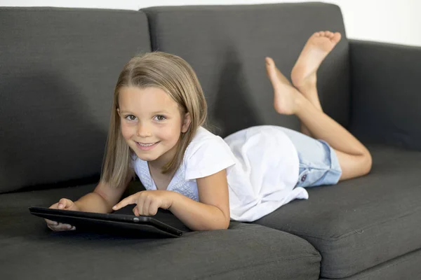 तरुण गोड आणि आनंदी लहान मुलगी इंटरनेट डिजिटल टॅब्लेट टच पॅड वापरून घर लिव्हिंग रूम सोफा सोफ्यावर 6 किंवा 7 वर्षांची झोपलेली — स्टॉक फोटो, इमेज