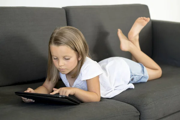 Jovem doce e feliz menina 6 ou 7 anos de idade deitado em casa sala de estar sofá usando internet tablet digital touch pad — Fotografia de Stock