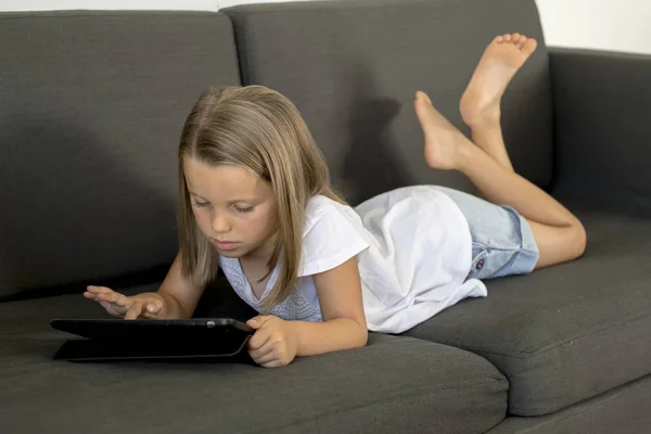 Jovem doce e feliz menina 6 ou 7 anos de idade deitado em casa sala de estar sofá usando internet tablet digital touch pad — Fotografia de Stock