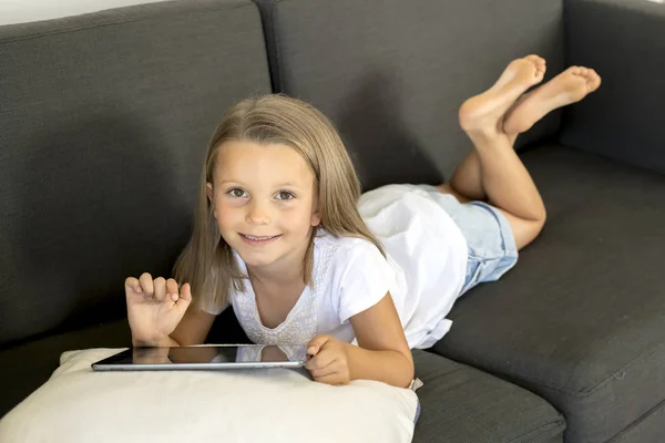 Jovem doce e feliz menina 6 ou 7 anos de idade deitado em casa sala de estar sofá usando internet tablet digital touch pad — Fotografia de Stock