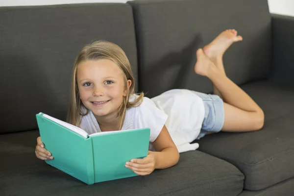 Young sweet and happy little girl 6 or 7 years old lying on home living room sofa couch reading a book quiet and adorable in children education — Stock Photo, Image