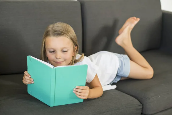 Young sweet and happy little girl 6 or 7 years old lying on home living room sofa couch reading a book quiet and adorable in children education — Stock Photo, Image