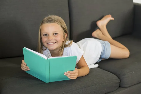 Unga söta och glada lilla tjej 6 eller 7 år gammal ligger hem vardagsrum soffa soffan en bok lugn och bedårande barn utbildning — Stockfoto