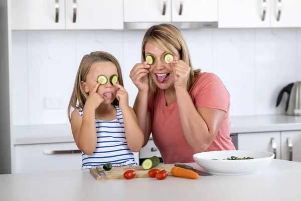有吸引力的妇女一起烹调与她甜美美丽的金发小6或7岁的女儿快乐嬉戏与黄瓜作为眼睛 — 图库照片