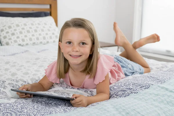 Doce e bonita loira 6 ou 7 anos menina deitada na cama sorrindo feliz usando a internet no tablet digital pad assistindo e se divertindo — Fotografia de Stock