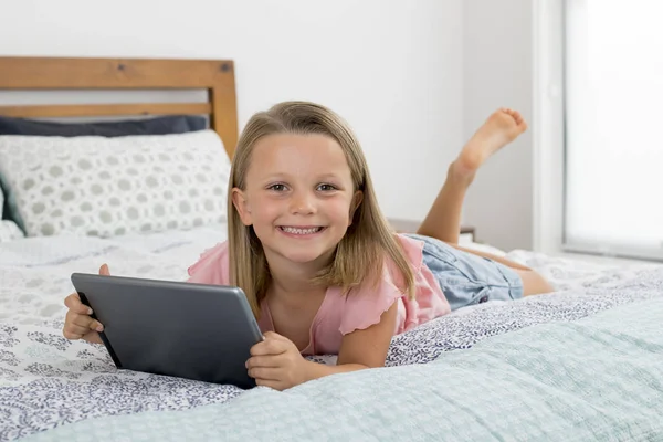 Doce e bonita loira 6 ou 7 anos menina deitada na cama sorrindo feliz usando a internet no tablet digital pad assistindo e se divertindo — Fotografia de Stock