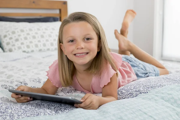 Сладкая и красивая блондинка 6 или 7 лет молодая девушка лежит на кровати улыбаясь счастливый с помощью Интернета на цифровом планшете смотреть и получать удовольствие — стоковое фото