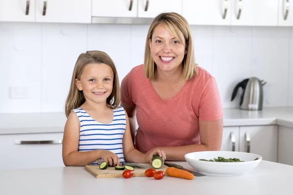 Joven atractiva mujer cocina junto con su dulce hermosa rubia poco 6 o 7 años hija sonriendo feliz preparando ensalada — Foto de Stock