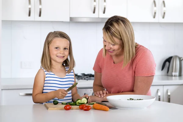 Jovem mulher atraente cozinhar junto com sua doce bela loira pequena 6 ou 7 anos de idade filha sorrindo feliz preparando salada — Fotografia de Stock