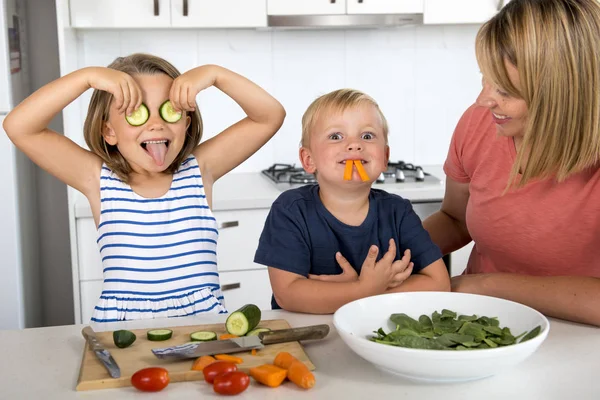 Joven atractiva mujer cocina junto con poco 3 y 7 años de edad hijo e hija jugando feliz con la barra de pepino como ojos preparando ensalada — Foto de Stock