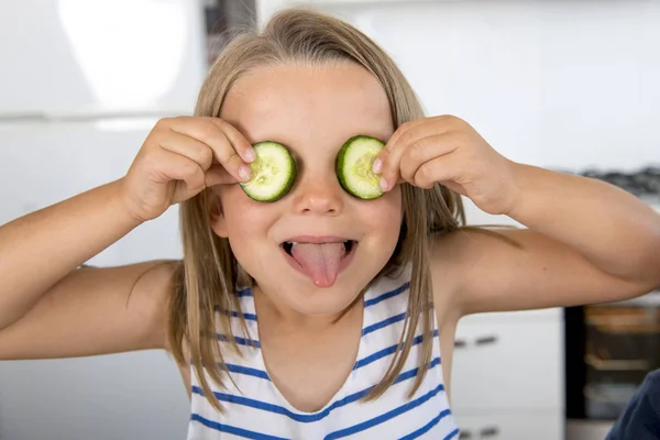 Muda cantik dan menggemaskan gadis 6 atau 7 tahun bersenang-senang di dapur rumah menempatkan potongan mentimun di matanya mengambil lidah bahagia — Stok Foto