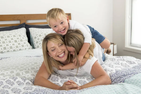 Jovem loira caucasiana mulher deitada na cama, juntamente com seu pequeno doce 3 e 7 anos de idade filho e filha rindo jogando feliz em casa — Fotografia de Stock