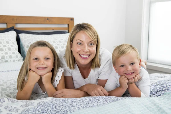 Jovem loira caucasiano mulher deitada na cama, juntamente com seu pequeno doce 3 e 7 anos de idade filho e filha sorrindo brincalhão e feliz — Fotografia de Stock