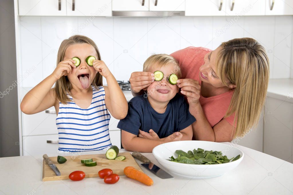 young attractive woman cooking together with little 3 and 7 years old son and daughter playing happy with cucumber rod as eyes preparing salad