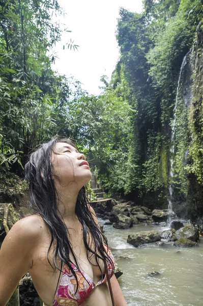 Portrait of young beautiful Asian girl looking pure and enjoying nature beauty with face wet under amazing beautiful natural waterfall in tropical paradise — Stock Photo, Image