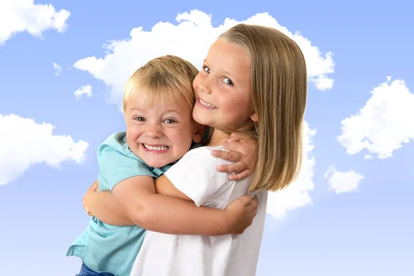 7 jaar oude schattige blonde gelukkig meisje poseren met haar kleine 3 jaar oude broer lacht vrolijk geïsoleerd op blauwe hemel met wolken — Stockfoto