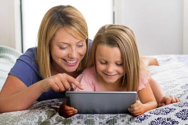 Loira caucasiano mãe deitado na cama com seu jovem doce 7 anos de idade filha usando internet no digital internet tablet pad juntos — Fotografia de Stock