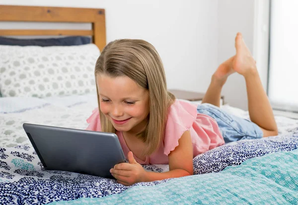 Bela loira 6 ou 7 anos de idade jovem deitada na cama sorrindo feliz usando a internet no tablet digital pad assistindo e se divertindo — Fotografia de Stock