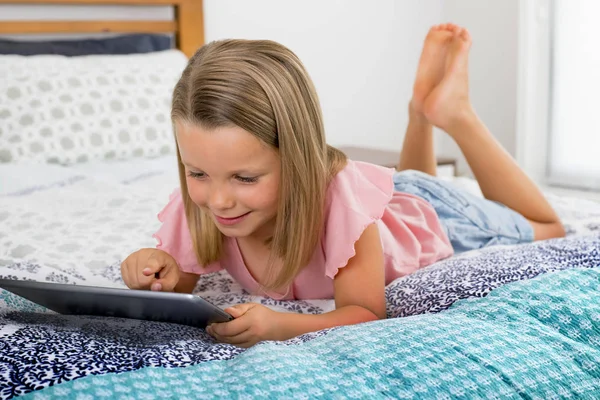 Красивая блондинка 6 или 7 лет молодая девушка лежит на кровати улыбаясь счастливый с помощью Интернета на цифровой планшет смотреть и получать удовольствие — стоковое фото