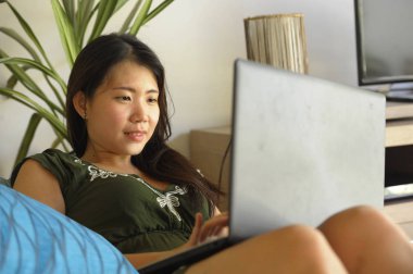 genç güzel ve rahat Asyalı Çinli kadın 20'li ya da 30'lu yaşlarda çalışma ya da bilgisayar dizüstü kullanma salon ev kanepe kanepede mutlu yalan