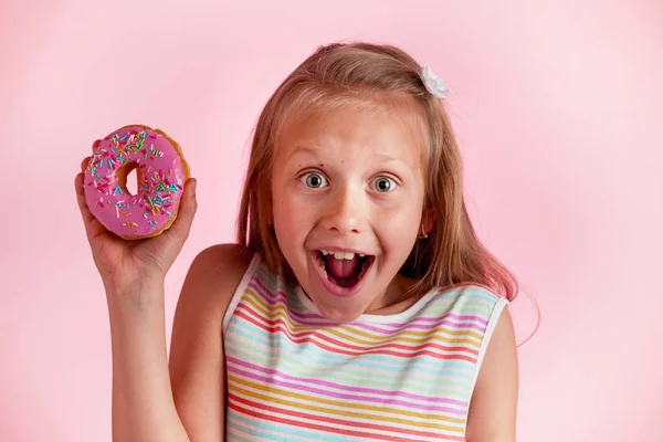 Cantik muda bahagia dan bersemangat gadis pirang 8 atau 9 tahun memegang donat di tangannya tampak kejang dan ceria dalam konsep kecanduan gula — Stok Foto