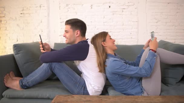 ソファ幸せ家庭でお互いを無視して携帯電話を使用してインターネット通信とスマート フォン中毒の概念の関係の欠如でアプリに集中して背中合わせに座っている若い魅力的なカップル — ストック動画