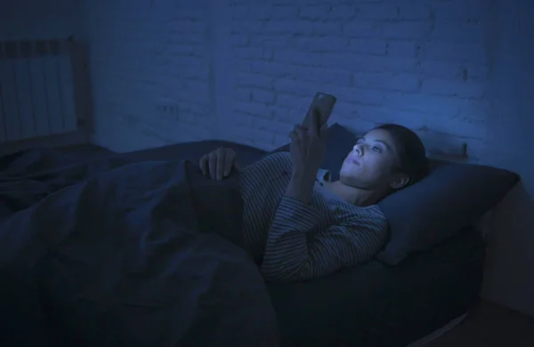 Retrato de la joven hermosa mujer latina utilizando el teléfono móvil tarde en la noche insomnio acostado en la cama en la oscuridad en el teléfono inteligente y la adicción a Internet citas aplicación Imagen de stock