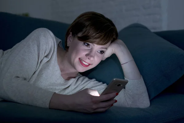 Красивая и счастливая рыжая женщина 20-30 лет лежит на домашнем диване или кровати с помощью мобильного телефона поздно ночью улыбаясь в интернет-зависимости — стоковое фото