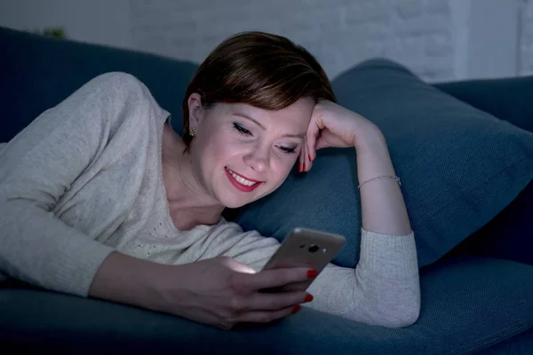 Mujer pelirroja bonita y feliz en sus 20 o 30 años acostada en el sofá o cama del hogar usando el teléfono móvil tarde en la noche sonriendo en la adicción a Internet —  Fotos de Stock