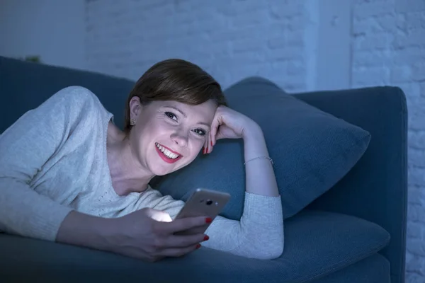 Mujer pelirroja bonita y feliz en sus 20 o 30 años acostada en el sofá o cama del hogar usando el teléfono móvil tarde en la noche sonriendo en la adicción a Internet —  Fotos de Stock