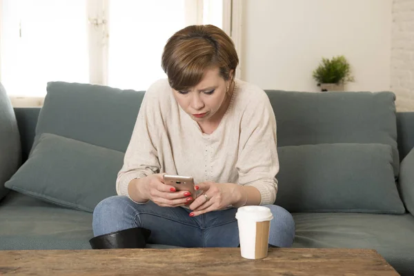 Joven atractiva mujer de 30 años de pelo rojo molesto aburrido y malhumorado utilizando aplicación de Internet en el teléfono móvil sentado en el sofá sofá de casa en la expresión de la cara molesta —  Fotos de Stock